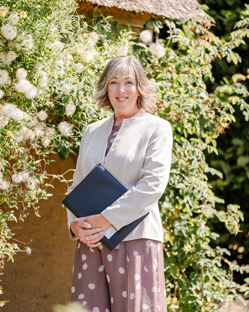 Veronica Hale smiling in a rose garden beside a thatched cottage, ceremony folder in hand