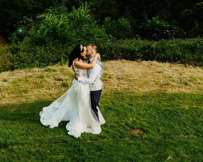 A bride and groom kissing on a grassy hillside, woodland behind them