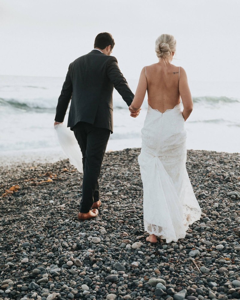 A couple walking barefoot along a pebble shoreline toward the sea