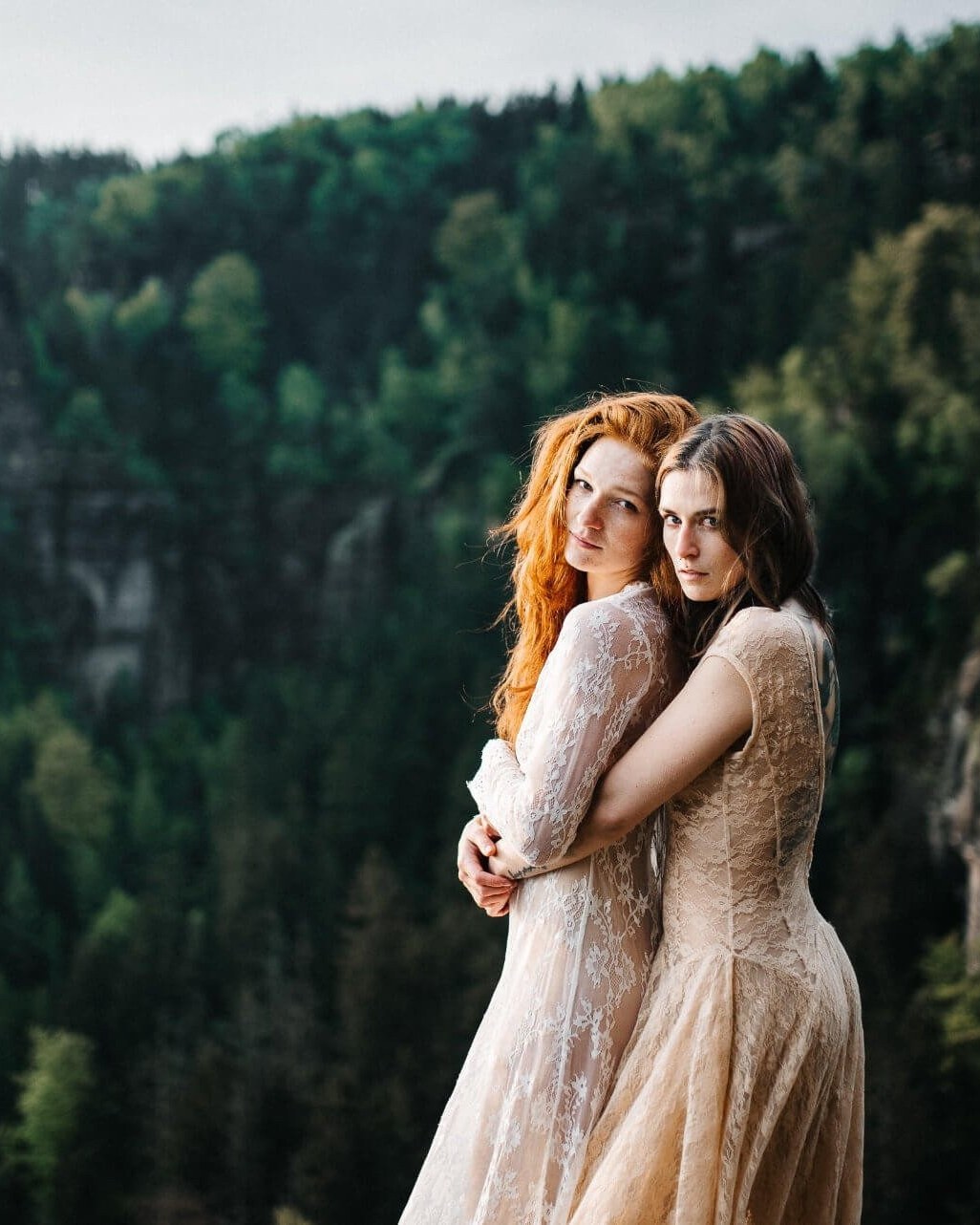 Two brides in lace holding each other on a clifftop above a wooded gorge